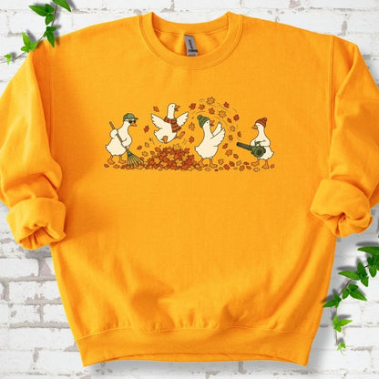 Gold sweatshirt flat lay with a row of four white cartoon geese doing fall yard work, one raking in a green cap and sunglasses, one leaping into a pile of orange and gold leaves in a plaid scarf, one in a green beanie tossing leaves and one in an orange beanie using a leaf blower, on white brick with ivy sprigs.