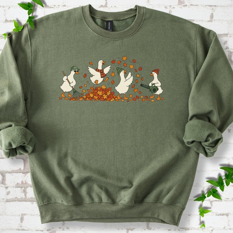 Military Green sweatshirt flat lay with a row of four white cartoon geese doing fall yard work, one raking in a green cap and sunglasses, one leaping into a pile of orange and gold leaves in a plaid scarf, one in a green beanie tossing leaves and one in an orange beanie using a leaf blower, on white brick with ivy sprigs.