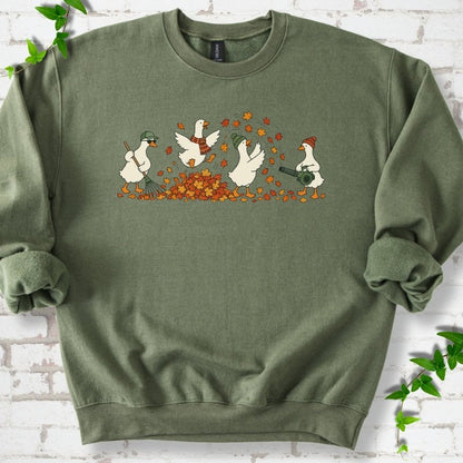 Military Green sweatshirt flat lay with a row of four white cartoon geese doing fall yard work, one raking in a green cap and sunglasses, one leaping into a pile of orange and gold leaves in a plaid scarf, one in a green beanie tossing leaves and one in an orange beanie using a leaf blower, on white brick with ivy sprigs.