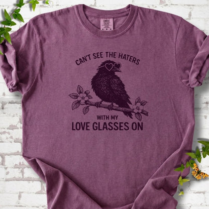 Berry T-shirt flat lay with deep plum "Can't see the haters with my love glasses on" lettering around a crow in heart-shaped sunglasses perched on a blossom branch, on white brick with ivy sprigs.