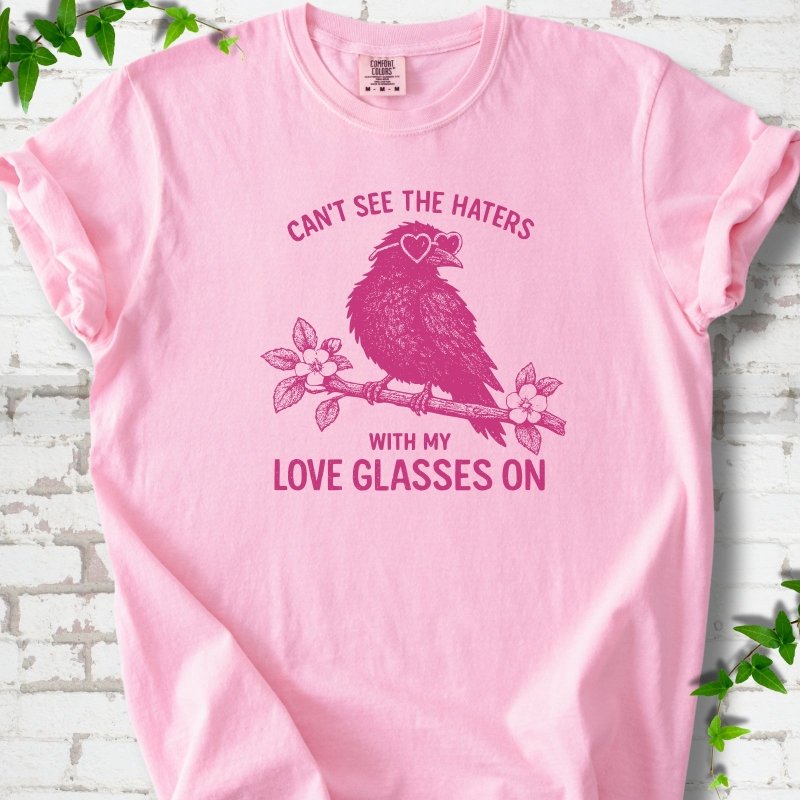 Blossom T-shirt flat lay with hot pink "Can't see the haters with my love glasses on" lettering around a crow in heart-shaped sunglasses perched on a blossom branch, on white brick with ivy sprigs.