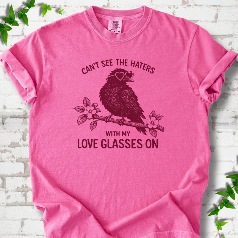 Crunchberry T-shirt flat lay with deep crimson "Can't see the haters with my love glasses on" lettering around a crow in heart-shaped sunglasses perched on a blossom branch, on white brick with ivy sprigs.