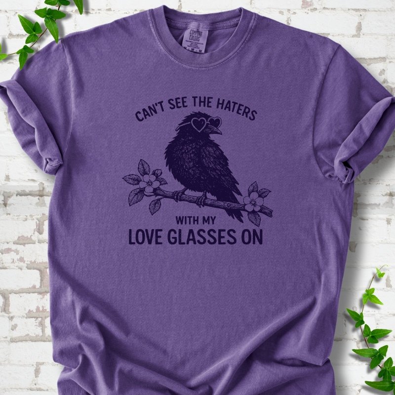 Grape T-shirt flat lay with deep indigo "Can't see the haters with my love glasses on" lettering around a crow in heart-shaped sunglasses perched on a blossom branch, on white brick with ivy sprigs.