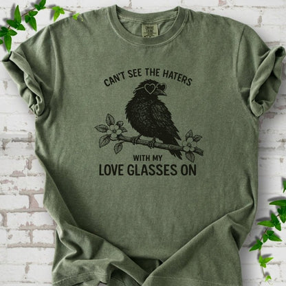 Hemp T-shirt flat lay with dark olive "Can't see the haters with my love glasses on" lettering around a crow in heart-shaped sunglasses perched on a blossom branch, on white brick with ivy sprigs.