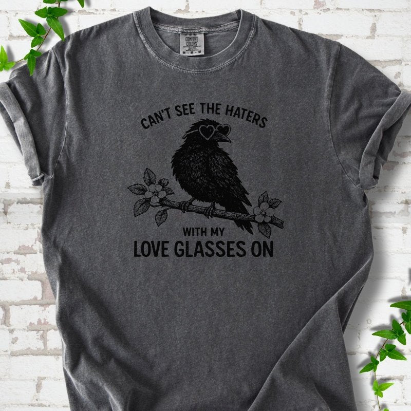 Pepper T-shirt flat lay with black "Can't see the haters with my love glasses on" lettering around a crow in heart-shaped sunglasses perched on a blossom branch, on white brick with ivy sprigs.