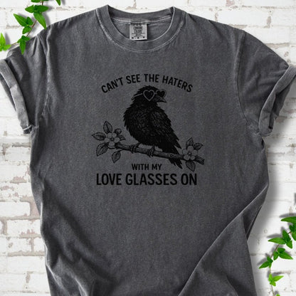 Pepper T-shirt flat lay with black "Can't see the haters with my love glasses on" lettering around a crow in heart-shaped sunglasses perched on a blossom branch, on white brick with ivy sprigs.
