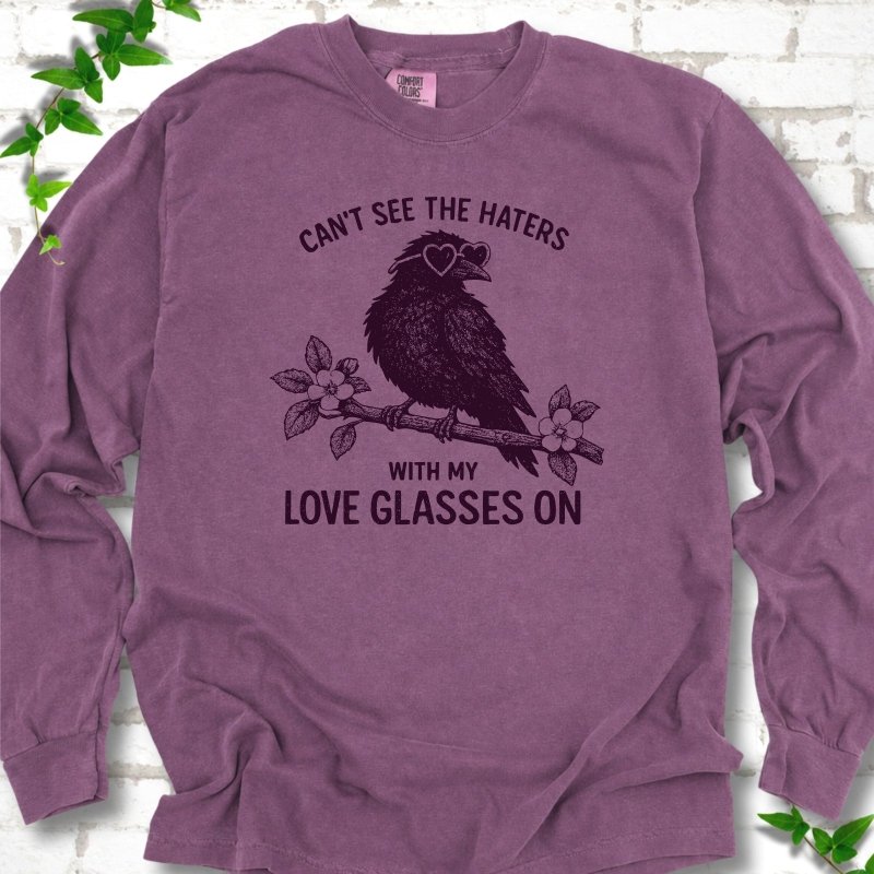 Berry long sleeve T-shirt flat lay with deep plum "Can't see the haters with my love glasses on" lettering around a crow in heart-shaped sunglasses perched on a blossom branch, on white brick with ivy sprigs.