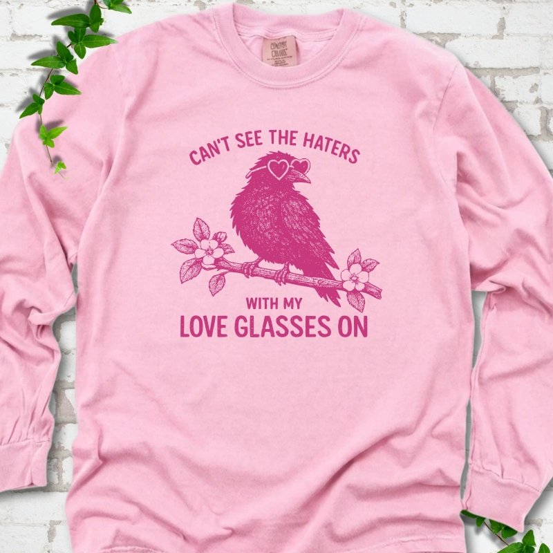 Blossom long sleeve T-shirt flat lay with hot pink "Can't see the haters with my love glasses on" lettering around a crow in heart-shaped sunglasses perched on a blossom branch, on white brick with ivy sprigs.