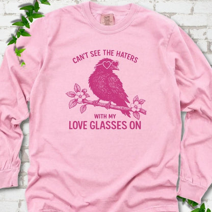 Blossom long sleeve T-shirt flat lay with hot pink "Can't see the haters with my love glasses on" lettering around a crow in heart-shaped sunglasses perched on a blossom branch, on white brick with ivy sprigs.