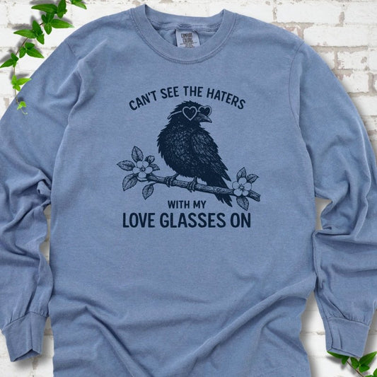 Blue Jean long sleeve T-shirt flat lay with navy "Can't see the haters with my love glasses on" lettering around a crow in heart-shaped sunglasses perched on a blossom branch, on white brick with ivy sprigs.