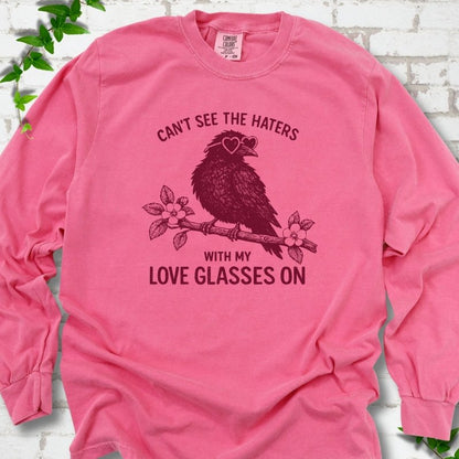 Crunchberry long sleeve T-shirt flat lay with deep crimson "Can't see the haters with my love glasses on" lettering around a crow in heart-shaped sunglasses perched on a blossom branch, on white brick with ivy sprigs.