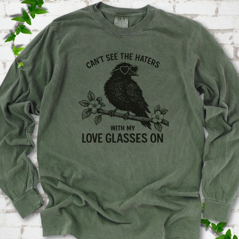 Hemp long sleeve T-shirt flat lay with dark olive "Can't see the haters with my love glasses on" lettering around a crow in heart-shaped sunglasses perched on a blossom branch, on white brick with ivy sprigs.