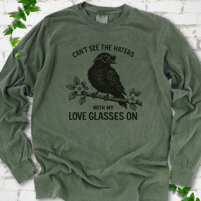 Hemp long sleeve T-shirt flat lay with dark olive "Can't see the haters with my love glasses on" lettering around a crow in heart-shaped sunglasses perched on a blossom branch, on white brick with ivy sprigs.