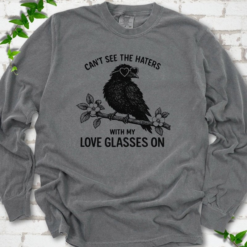 Pepper long sleeve T-shirt flat lay with black "Can't see the haters with my love glasses on" lettering around a crow in heart-shaped sunglasses perched on a blossom branch, on white brick with ivy sprigs.