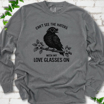 Pepper long sleeve T-shirt flat lay with black "Can't see the haters with my love glasses on" lettering around a crow in heart-shaped sunglasses perched on a blossom branch, on white brick with ivy sprigs.