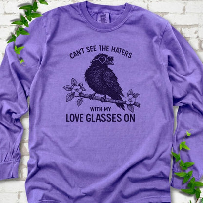 Violet long sleeve T-shirt flat lay with deep indigo "Can't see the haters with my love glasses on" lettering around a crow in heart-shaped sunglasses perched on a blossom branch, on white brick with ivy sprigs.