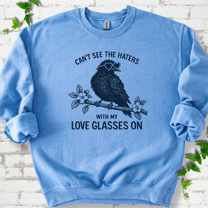 Carolina Blue sweatshirt flat lay with navy "Can't see the haters with my love glasses on" lettering around a crow in heart-shaped sunglasses perched on a blossom branch, on white brick with ivy sprigs.