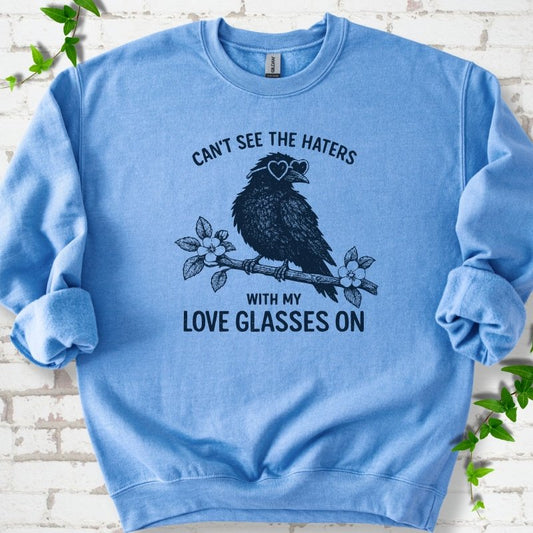 Carolina Blue sweatshirt flat lay with navy "Can't see the haters with my love glasses on" lettering around a crow in heart-shaped sunglasses perched on a blossom branch, on white brick with ivy sprigs.
