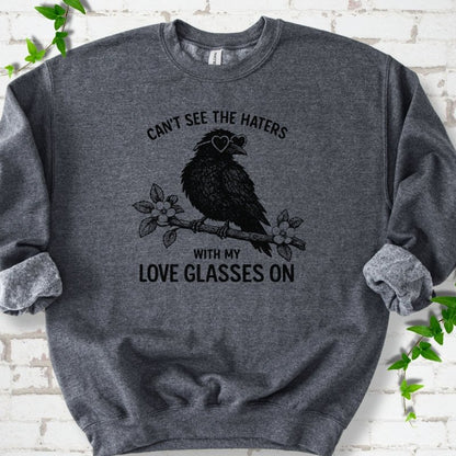 Dark Heather sweatshirt flat lay with black "Can't see the haters with my love glasses on" lettering around a crow in heart-shaped sunglasses perched on a blossom branch, on white brick with ivy sprigs.