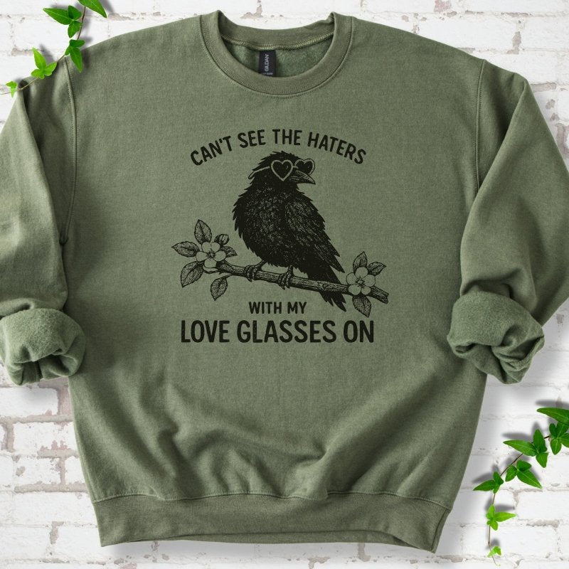 Military Green sweatshirt flat lay with dark olive "Can't see the haters with my love glasses on" lettering around a crow in heart-shaped sunglasses perched on a blossom branch, on white brick with ivy sprigs.
