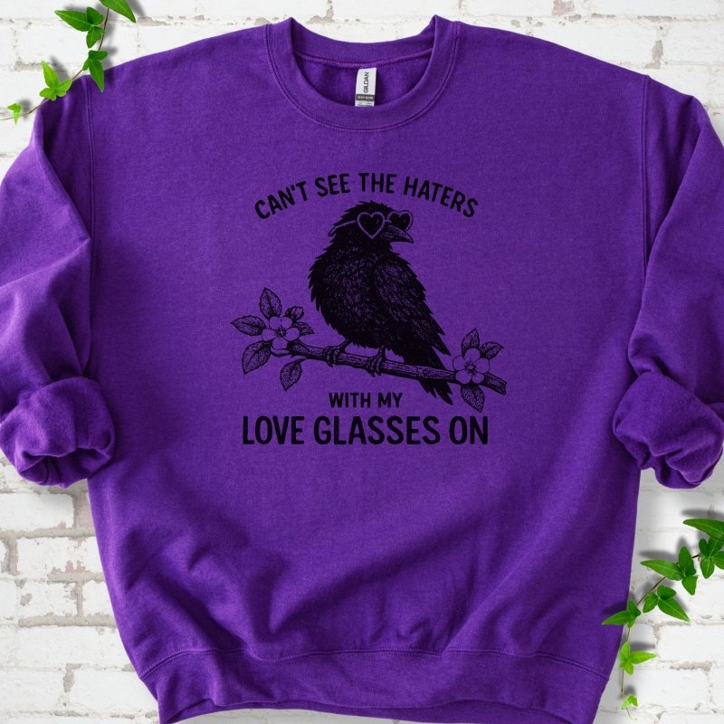 Purple sweatshirt flat lay with black "Can't see the haters with my love glasses on" lettering around a crow in heart-shaped sunglasses perched on a blossom branch, on white brick with ivy sprigs.