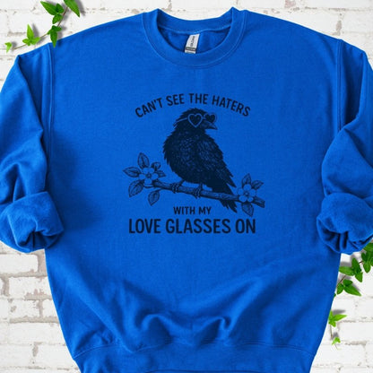 Royal sweatshirt flat lay with navy "Can't see the haters with my love glasses on" lettering around a crow in heart-shaped sunglasses perched on a blossom branch, on white brick with ivy sprigs.