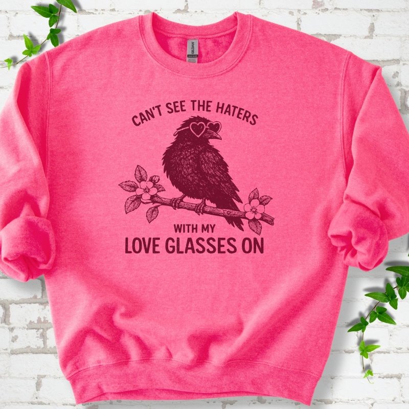 Safety Pink sweatshirt flat lay with deep crimson "Can't see the haters with my love glasses on" lettering around a crow in heart-shaped sunglasses perched on a blossom branch, on white brick with ivy sprigs.