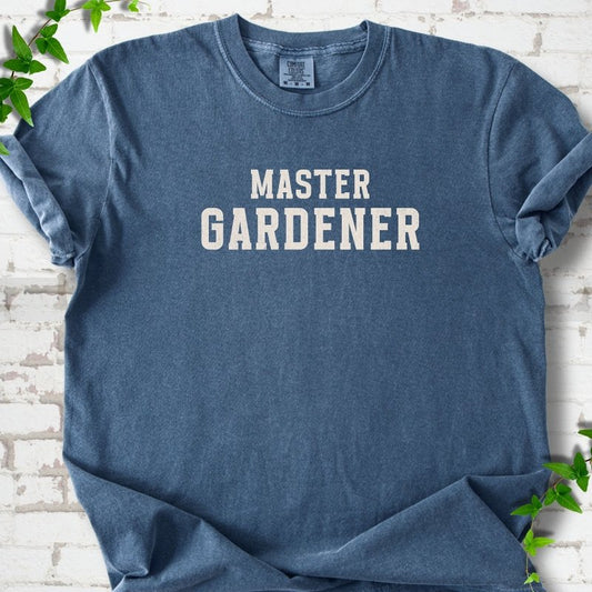 Denim short sleeve T-shirt laid flat on white brick with greenery, showing the cream words 'Master Gardener' centered on the chest.