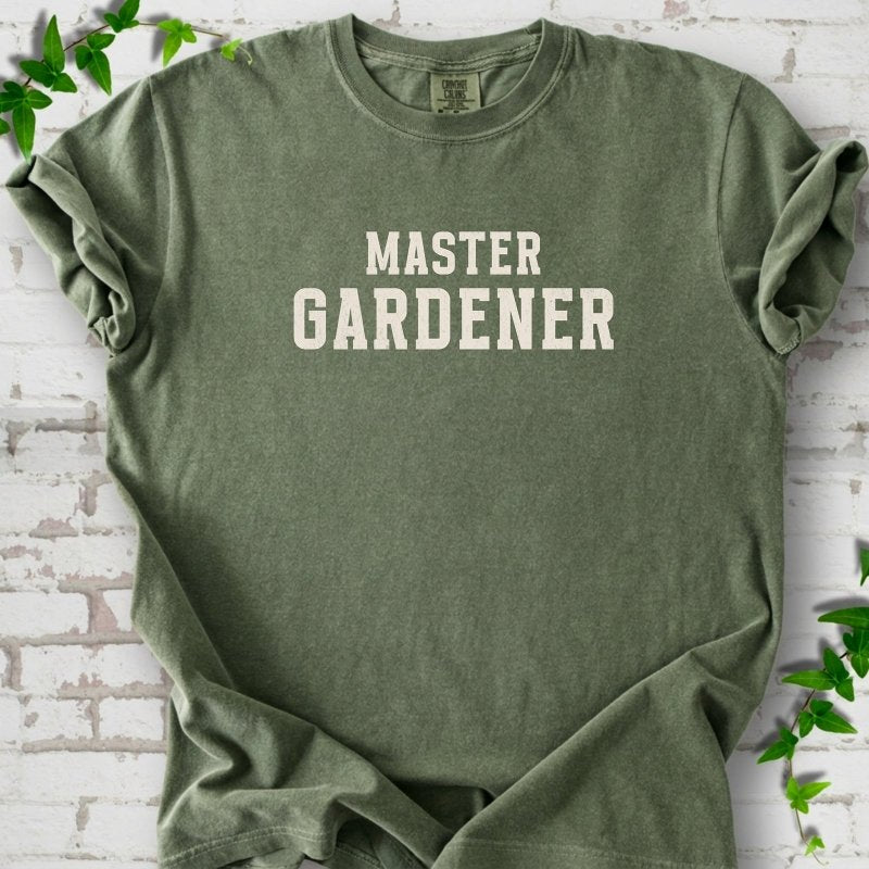 Moss short sleeve T-shirt laid flat on white brick with greenery, showing the cream words 'Master Gardener' centered on the chest.