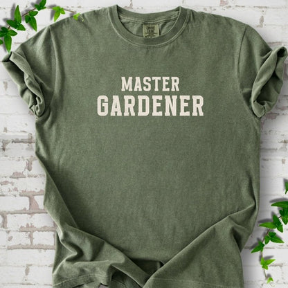Moss short sleeve T-shirt laid flat on white brick with greenery, showing the cream words 'Master Gardener' centered on the chest.