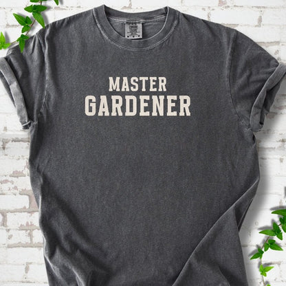 Pepper short sleeve T-shirt laid flat on white brick with greenery, showing the cream words 'Master Gardener' centered on the chest.
