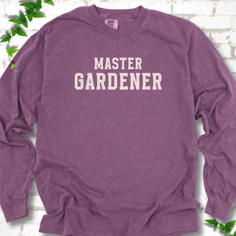 Berry long sleeve T-shirt laid flat on white brick with greenery, showing the cream words 'Master Gardener' centered on the chest.