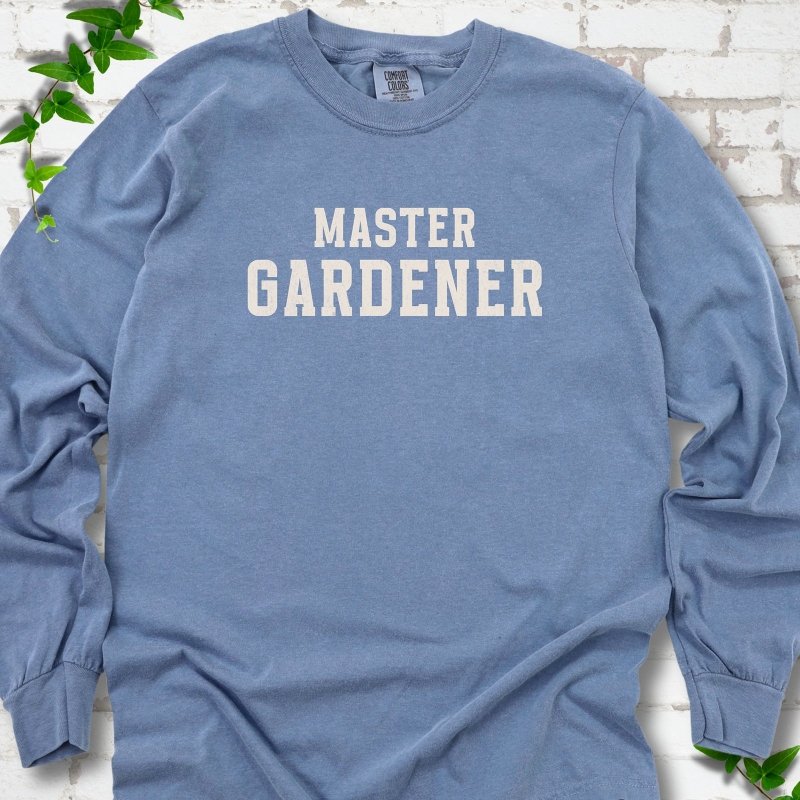 Blue Jean long sleeve T-shirt laid flat on white brick with greenery, showing the cream words 'Master Gardener' centered on the chest.