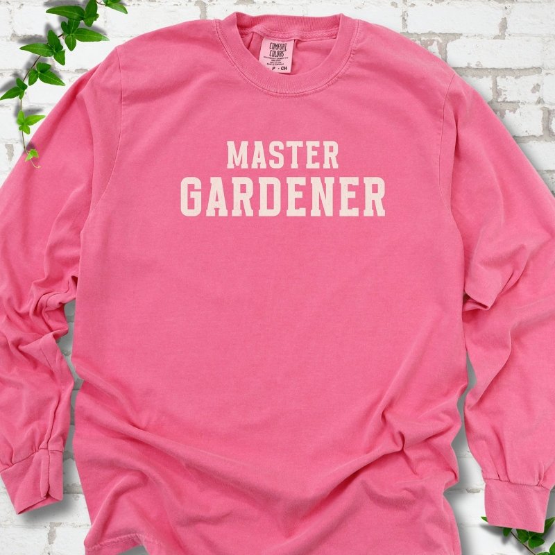 Crunchberry long sleeve T-shirt laid flat on white brick with greenery, showing the cream words 'Master Gardener' centered on the chest.