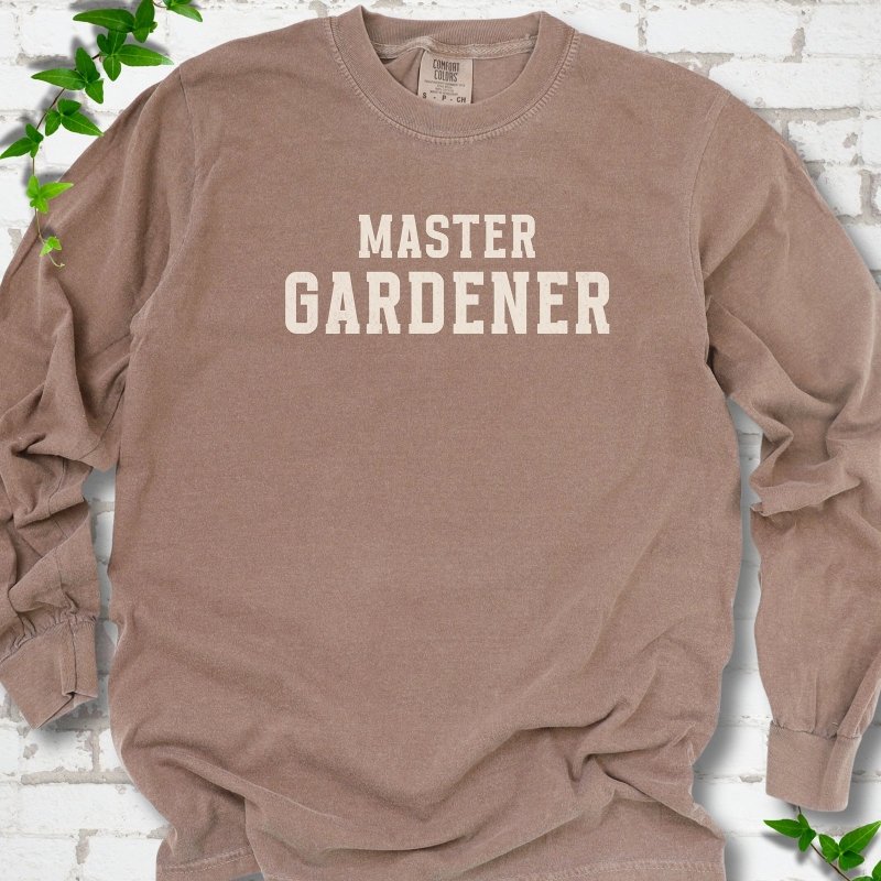 Espresso long sleeve T-shirt laid flat on white brick with greenery, showing the cream words 'Master Gardener' centered on the chest.