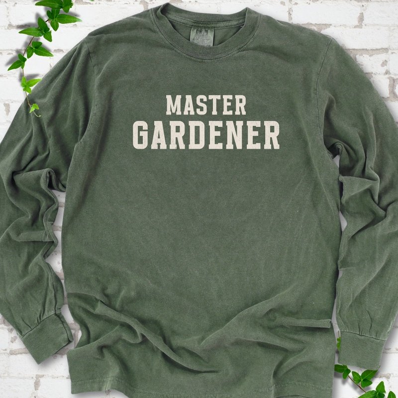 Hemp long sleeve T-shirt laid flat on white brick with greenery, showing the cream words 'Master Gardener' centered on the chest.