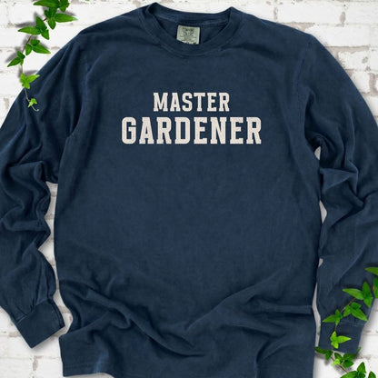 Midnight long sleeve T-shirt laid flat on white brick with greenery, showing the cream words 'Master Gardener' centered on the chest.