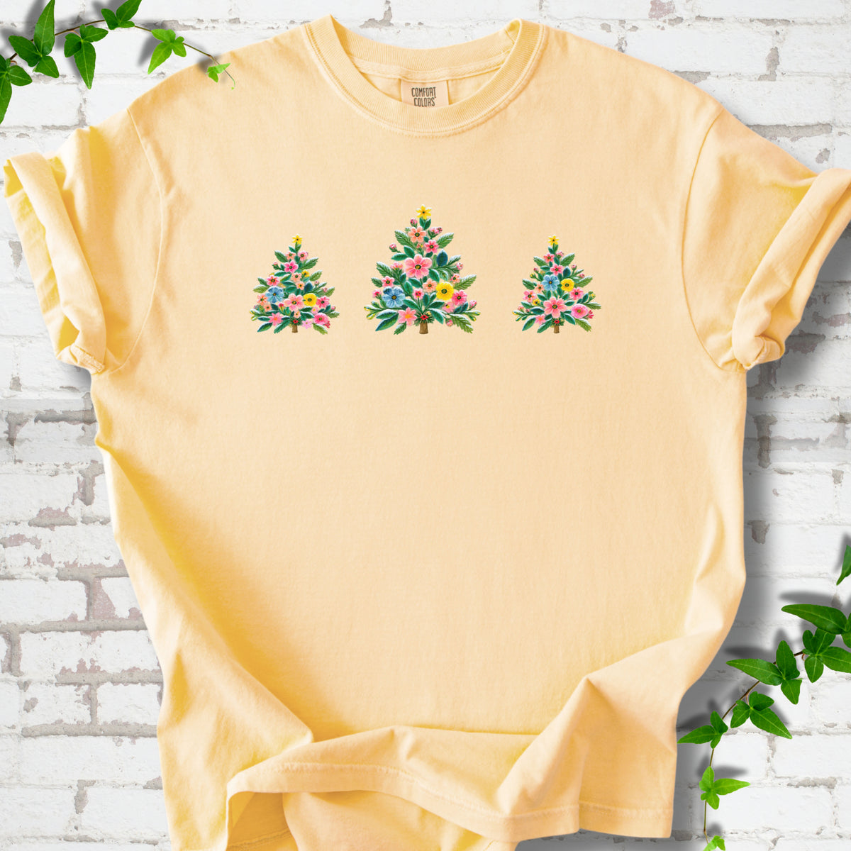 Butter short-sleeve T-shirt laid flat on white brick with three flower-filled trees centered on the front.
