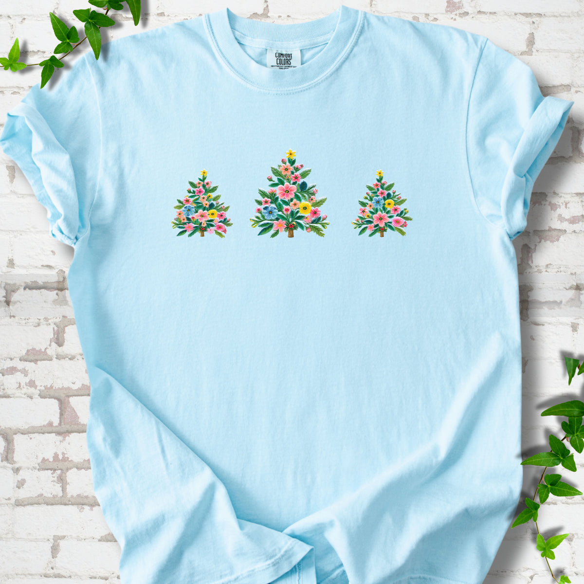 Chambray short-sleeve T-shirt laid flat on white brick with three flower-filled trees centered on the front.