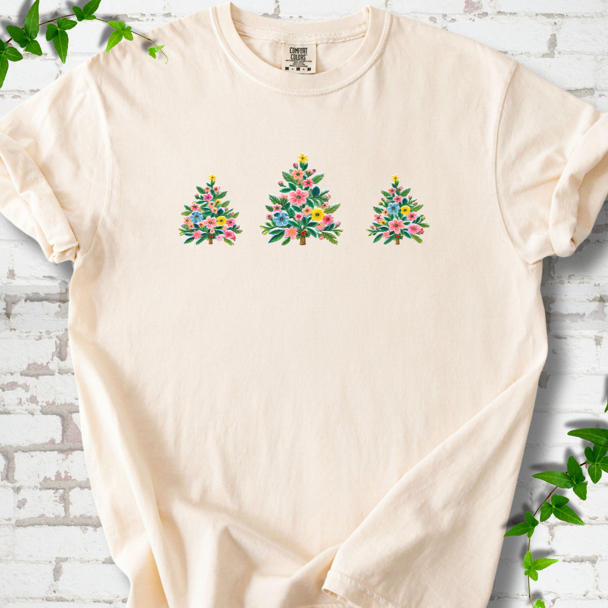 Ivory short-sleeve T-shirt laid flat on white brick with three flower-filled trees centered on the front.