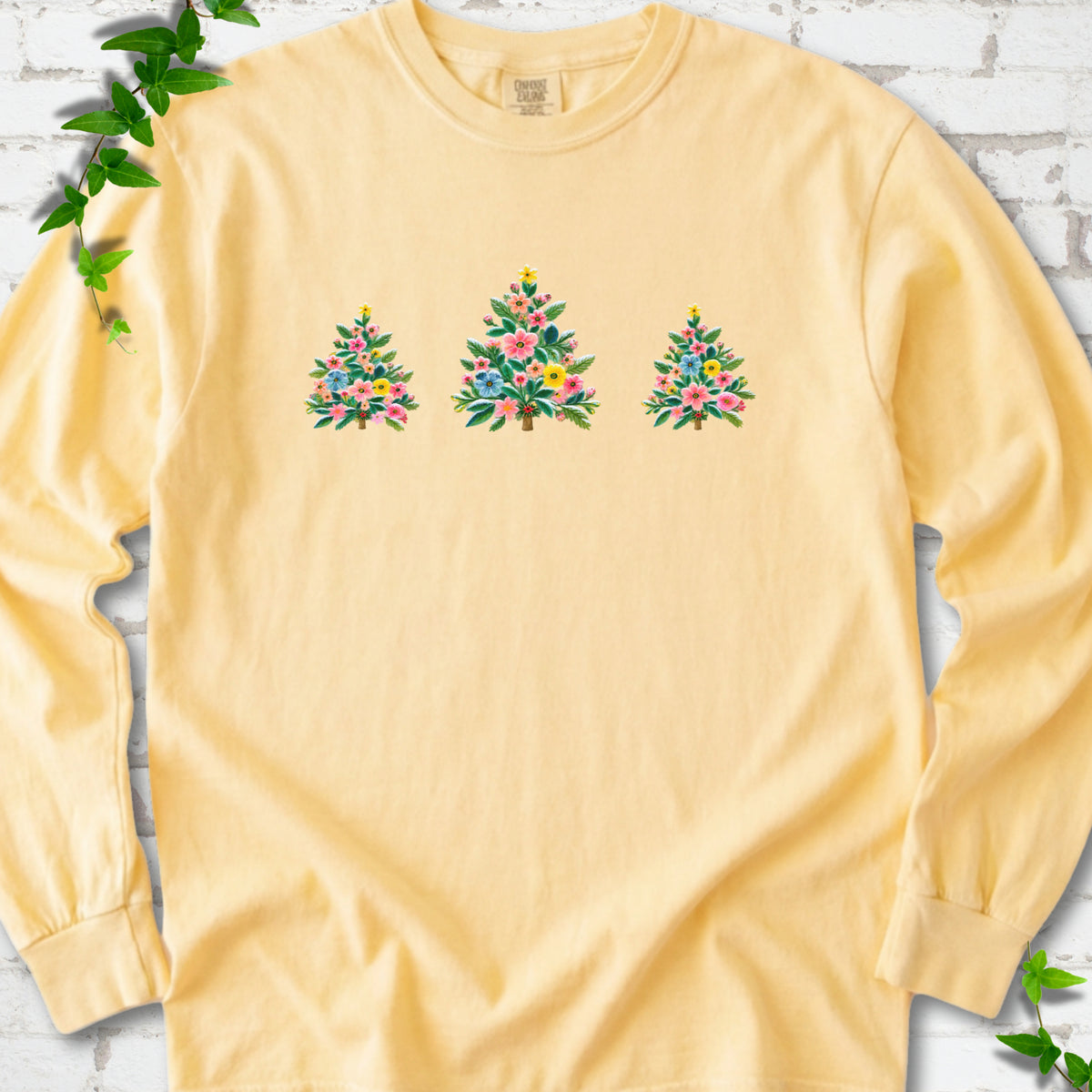Butter long-sleeve T-shirt laid flat on white brick with three flower-filled trees centered on the front.