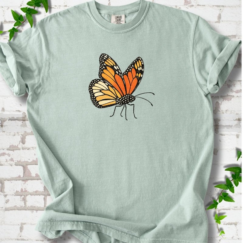 Bay light green short-sleeve T-shirt with one large orange and black monarch butterfly, wings raised, printed on the chest, flat lay on white brick with ivy