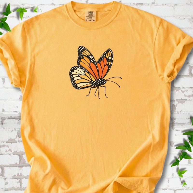 Citrus golden yellow short-sleeve T-shirt with one large orange and black monarch butterfly, wings raised, printed on the chest, flat lay on white brick with ivy