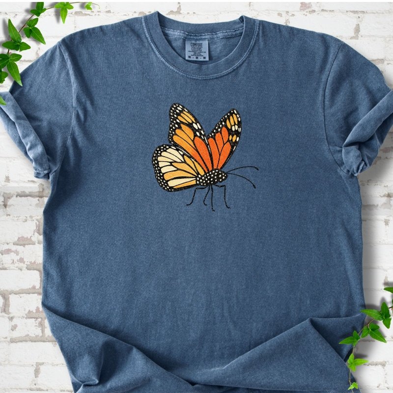 Denim blue short-sleeve T-shirt with one large orange and black monarch butterfly, wings raised, printed on the chest, flat lay on white brick with ivy