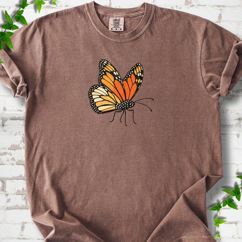Espresso brown short-sleeve T-shirt with one large orange and black monarch butterfly, wings raised, printed on the chest, flat lay on white brick with ivy