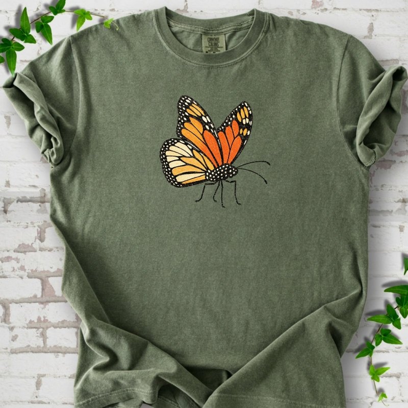 Moss green short-sleeve T-shirt with one large orange and black monarch butterfly, wings raised, printed on the chest, flat lay on white brick with ivy
