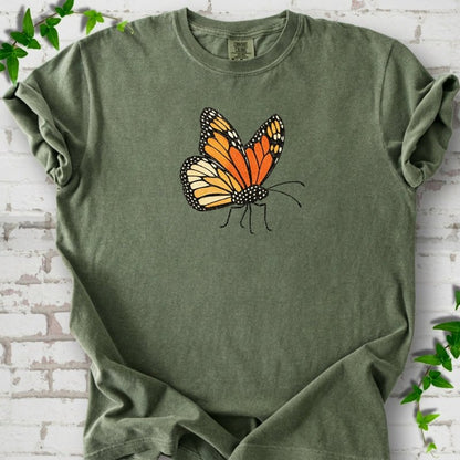 Moss green short-sleeve T-shirt with one large orange and black monarch butterfly, wings raised, printed on the chest, flat lay on white brick with ivy