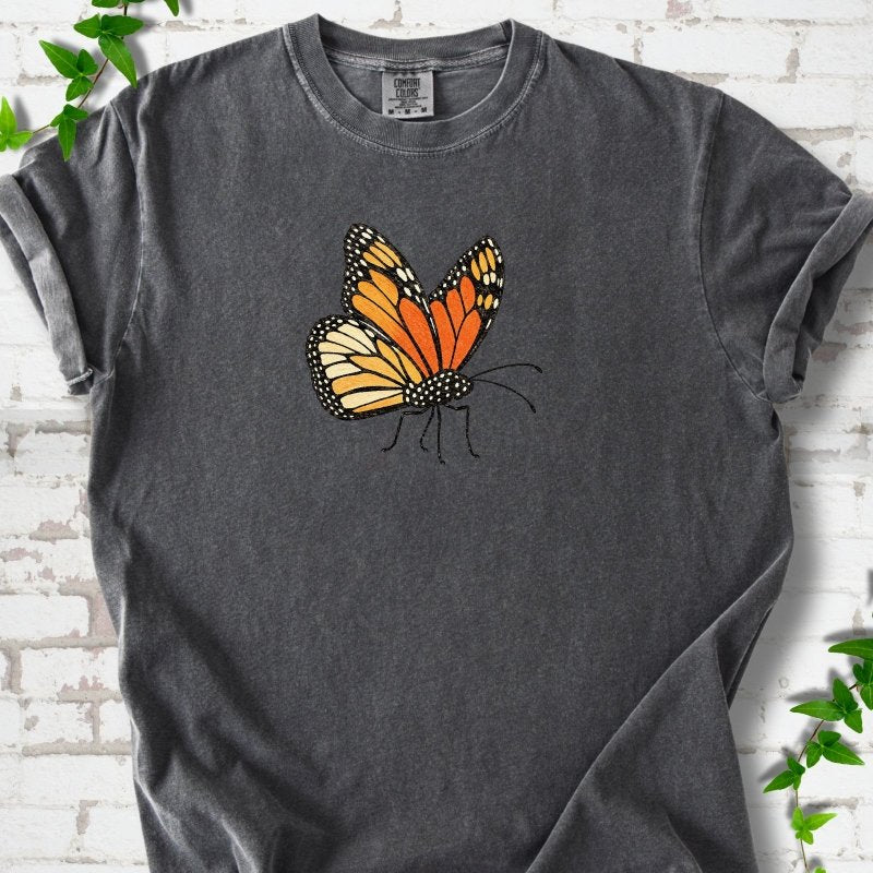 Pepper charcoal gray short-sleeve T-shirt with one large orange and black monarch butterfly, wings raised, printed on the chest, flat lay on white brick with ivy