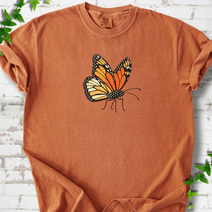 Yam burnt orange short-sleeve T-shirt with one large orange and black monarch butterfly, wings raised, printed on the chest, flat lay on white brick with ivy