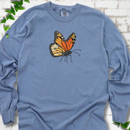 Blue Jean light blue long sleeve T-shirt with one large orange and black monarch butterfly, wings raised, printed on the chest, flat lay on white brick with ivy